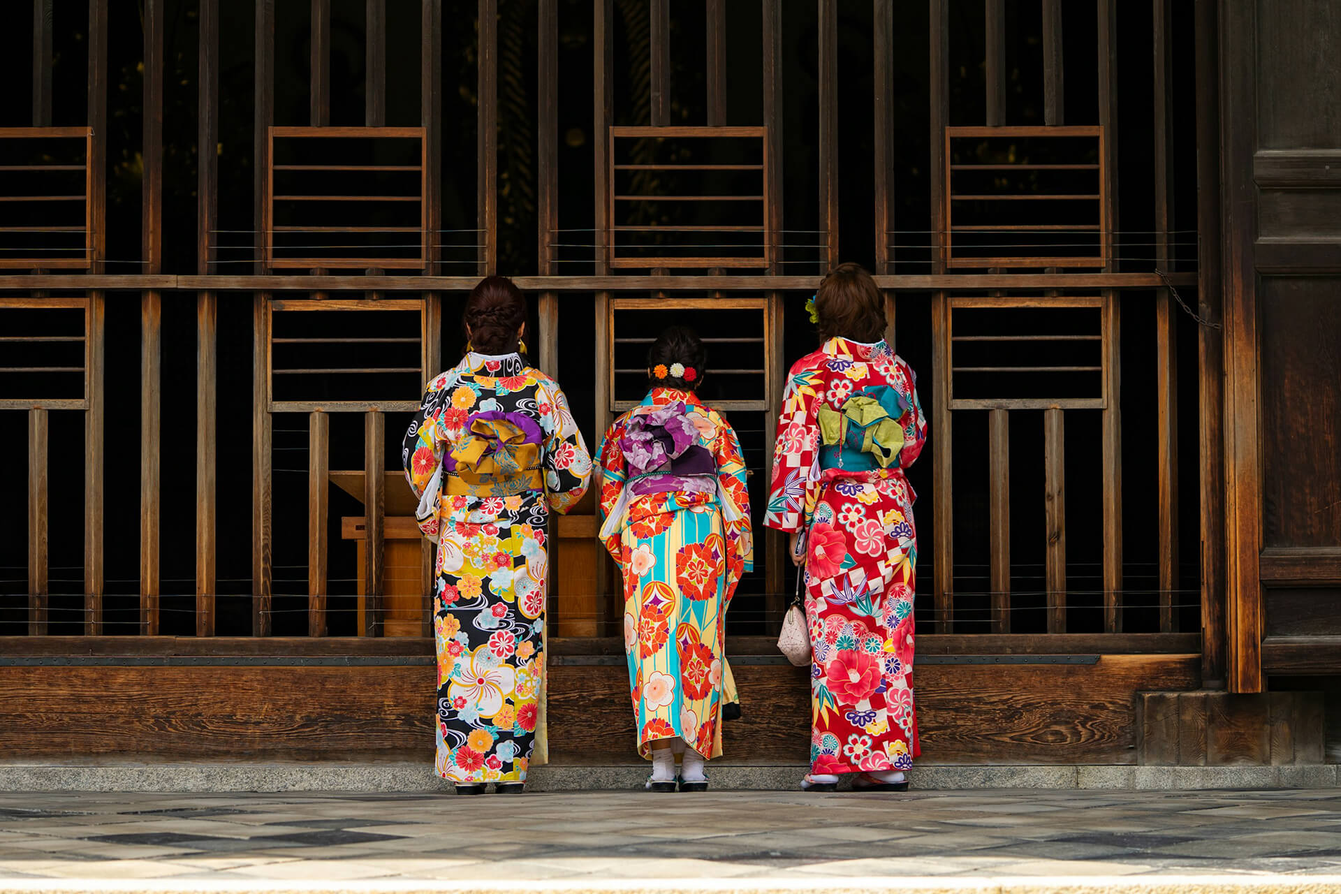 Three individuals in colorful kimonos stand with their backs to the camera, facing a traditional wooden gate. The kimonos feature floral patterns in vibrant hues, and each person carries an obi. The scene suggests a cultural or ceremonial setting.